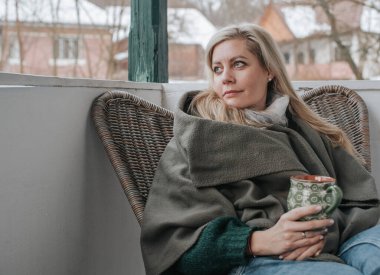 Sarışın bir kadın balkonda hasır bir sandalyede oturuyor, sıcak bir battaniyeye sarılı ve dekoratif bir kupa tutuyor, düşünceli bir şekilde uzaktan bakıyor.