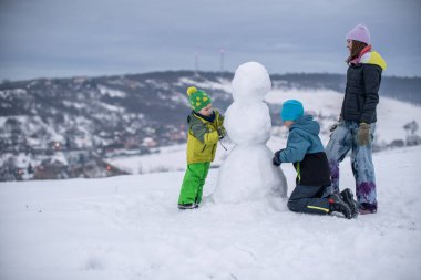 Üç çocuk etkin bir şekilde arka planda köyü olan karlı bir yamaçta kardan adam yapmakla meşguller.