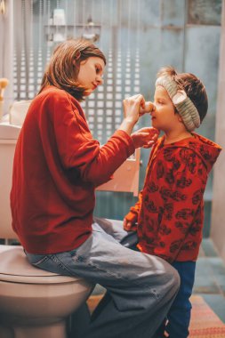 Genç bir kız klozet kapağına oturur ve küçük kardeşinin burnuna makyaj yapar.