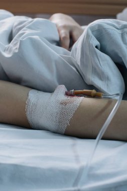 Hastanın kolunun yakından görüntüsü. Serum takılmış ve bandajlanmış. Hastane yatağında beyaz çarşafların altında yatıyor.