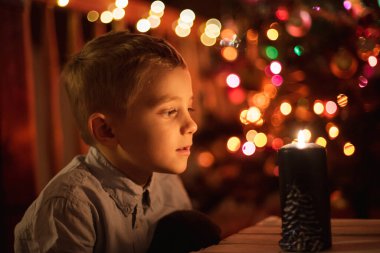 Yedi yaşında, arkasında mum ve Noel ağacı olan bir çocuk.