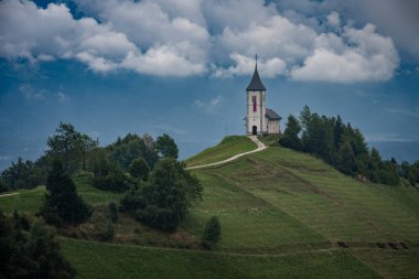 Slovenya 'nın Jamnik kentinde çakıl yollu dramatik bulutlu bir gökyüzünün altında manzaralı bir tepenin üzerinde duran İkonik Aziz Primus Kilisesi ve Felicianstanding,