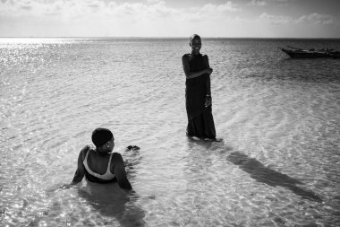 MICHAMVI, ZANZIBAR - 15 Ekim 2025: Güneşli bir günde sığ, ışıl ışıl okyanus suyunda dinlenen ve yürüyen iki Afrikalı kadının siyah-beyaz yaşam tarzı fotoğrafı.