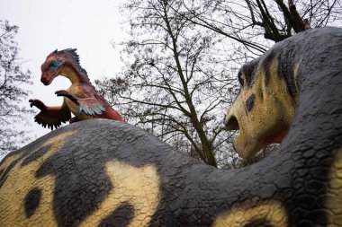 LUBIN, POLAND - 15 Ocak 2026 - Park Wroclawski. Küçük tüylü bir dinozor modeli, muhtemelen Deinonychus (Deinonychus), eğitici bir paleo parkında daha büyük bir dinozorun arkasına tünemiştir..