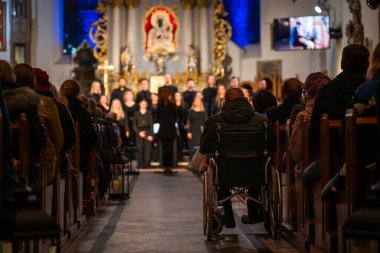 Interior of a church during a choir concert with audience in pews and a person in a wheelchair in the aisle, Poland.