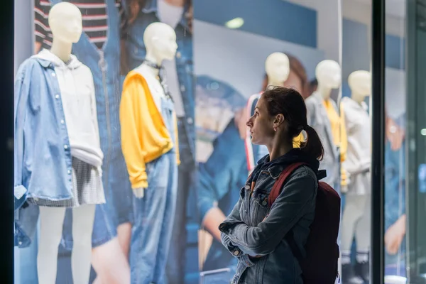 Girl stands outside the store and looks at the female clothes on the mannequins.