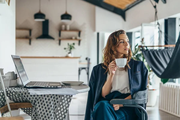 Une femme est assise dans la cuisine, distraite de travailler avec son ordinateur portable, buvant dans une tasse et regardant pensivement loin — Photo