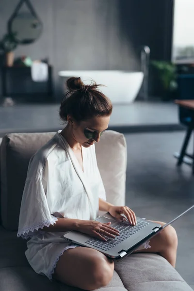 Woman sits at home on the couch in a nightgown with a laptop — Stock Photo, Image