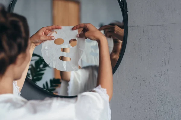 Vrouw met laken masker in haar handen in de badkamer voor het aanbrengen van het op haar gezicht — Stockfoto