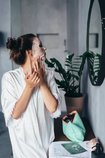 Kvinna applicerar sheet mask på hennes ansikte i badrummet — Stockfoto