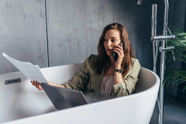 Žena sedí ve vaně s papíry, notebookem a telefonuje — Stock fotografie