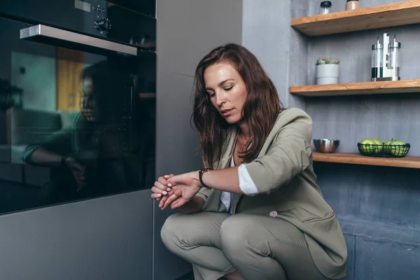 Mujer espera mirando el reloj para una comida mientras se sienta en el horno —  Fotos de Stock