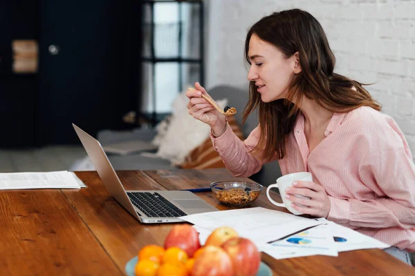 Egy fiatal nő eszik és nézi a laptopját. — Stock Fotó