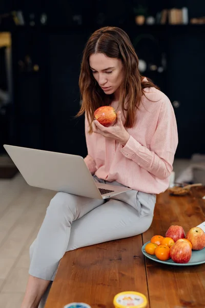Menina senta-se na mesa com laptop e come maçã — Fotografia de Stock