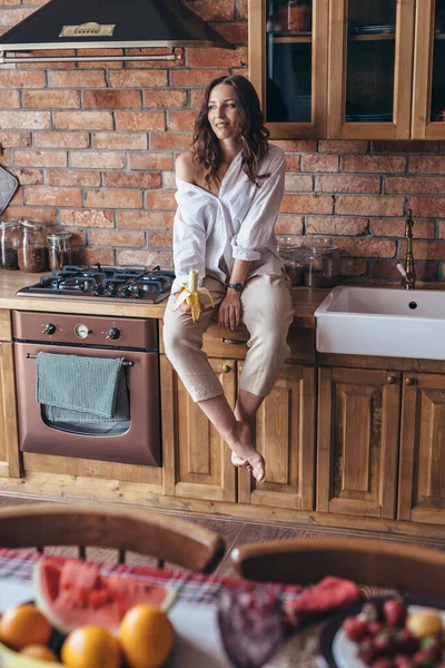 Jeune femme assise avec une banane dans la cuisine sur la table — Photo