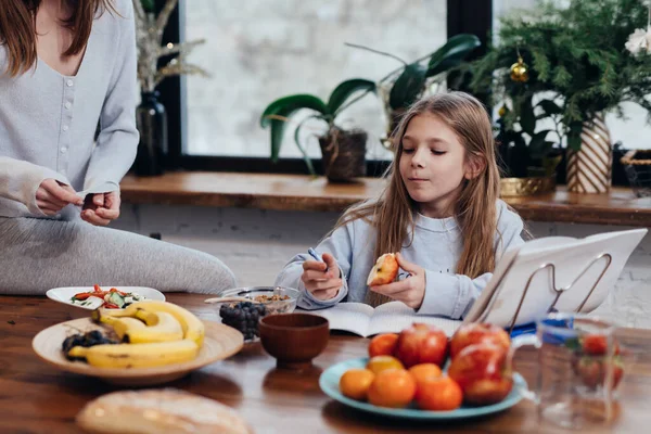 Chica hace su tarea en la cocina mientras su madre cocina. —  Fotos de Stock