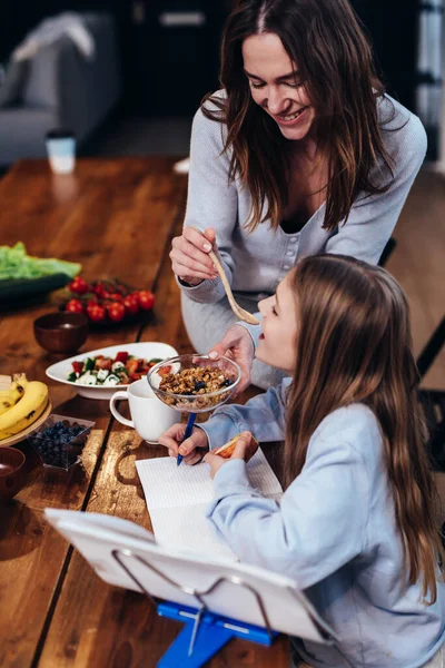 Flickan gör sina läxor vid köksbordet, och hennes mamma ger henne en smak av mat med en sked — Stockfoto