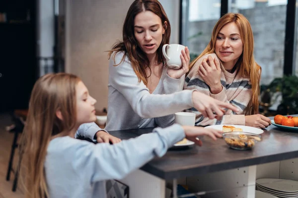 Freundinnen und ein kleines Mädchen sitzen zusammen am Küchentisch, trinken Tee und unterhalten sich. — Stockfoto