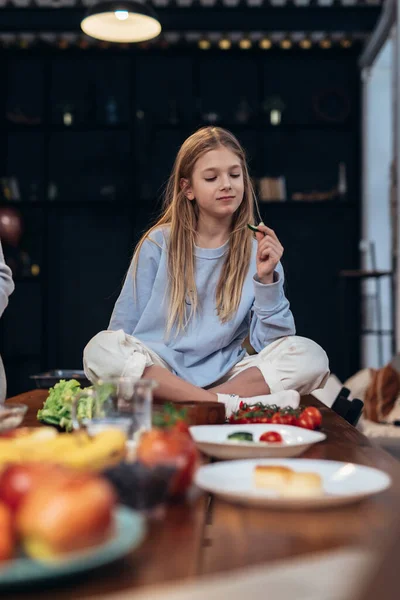 Flicka äter grönsaker sittande på köksbordet. — Stockfoto