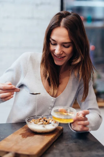 Ung kvinna i köket tillför honung till müsli. — Stockfoto