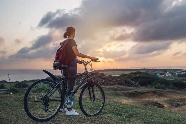 Young woman watching the sunset and the beautiful view of nature while standing with her bicycle on a hill. — Stock Photo, Image