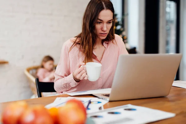 Jovem mulher trabalha na cozinha em seu laptop enquanto sua filha fica em casa — Fotografia de Stock