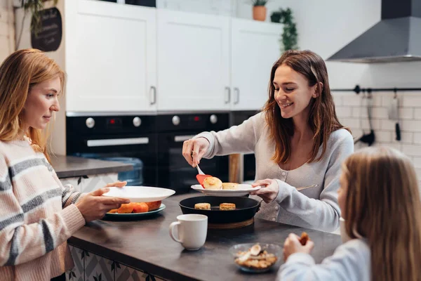 Young woman puts pancakes from the frying pan to her friend and daughter — Stock Photo, Image
