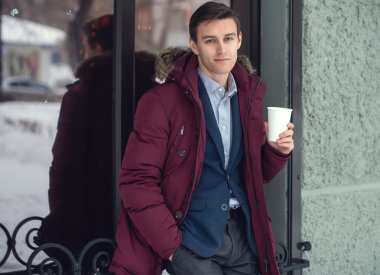 Young man in winter coat woth coffee to go standing against the cafe wall clipart