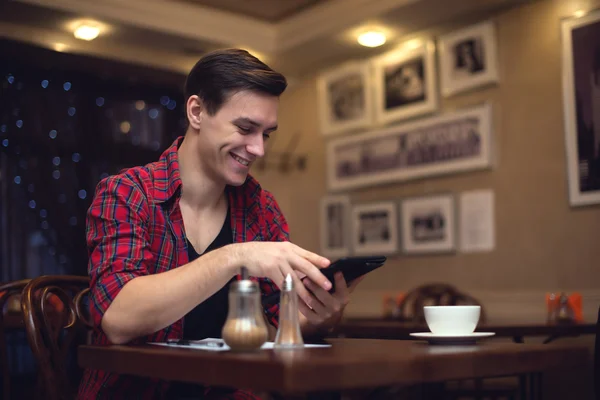 Joven sonriente atractivo hombre utiliza su tableta en la hora del almuerzo en la cafetería —  Fotos de Stock