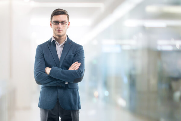 Young handsome businessman wearing glasses in the office and standing alone hands crossed.