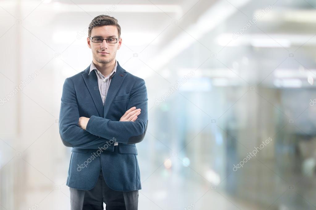 Young handsome businessman wearing glasses in the office and standing alone hands crossed.