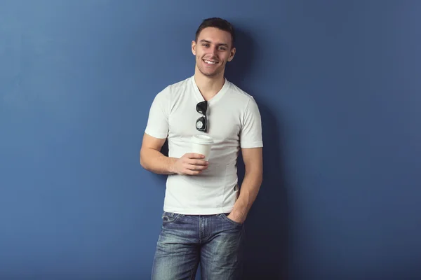 Mladý muž v šortky a bílé tričko se usmívá stojící u zdi s brýlemi kávy. — Stock fotografie