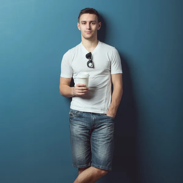 Joven atractivo hombre guapo en camiseta blanca pantalones vaqueros pantalones cortos de mezclilla de pie contra la pared y sonriendo sosteniendo café . — Foto de Stock