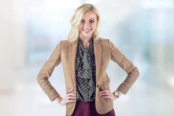 Jasny młoda kobieta w brązowy płaszcz jest stałego biura z ręce na jej biodrach i uśmiech jasne. — Zdjęcie stockowe