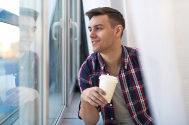 Handsome smiling man guy in plaid shirt looking through the window with a cup of morning coffee concept waiting dreaming