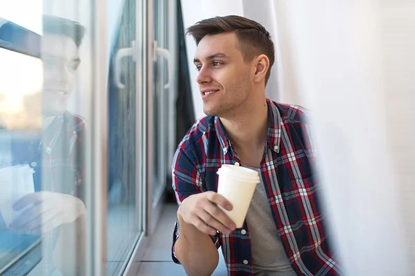 Guapo hombre sonriente en camisa a cuadros mirando a través de la ventana con una taza de café de la mañana concepto de espera soñando —  Fotos de Stock