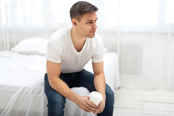 Hombre guapo en camiseta blanca y vaqueros azules sentado en la cama con una taza de café de la mañana . —  Fotos de Stock