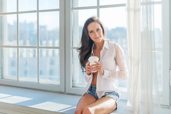 Hermosa chica vistiendo ropa sexy en casa sentada junto a la ventana mañana con una taza de café en sus manos llenas de luz —  Fotos de Stock