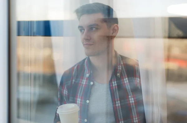 Guapo hombre sonriente en camisa a cuadros mirando a través de la ventana con una taza de café de la mañana concepto de espera soñando —  Fotos de Stock