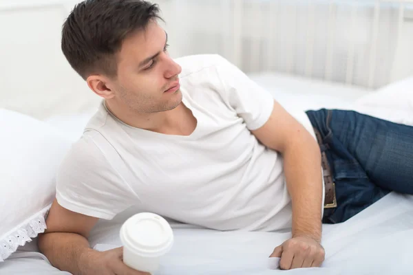 Красивый мужчина в белой футболке и синих джинсах лежит на кровати дома с чашкой утреннего кофе или чая — стоковое фото