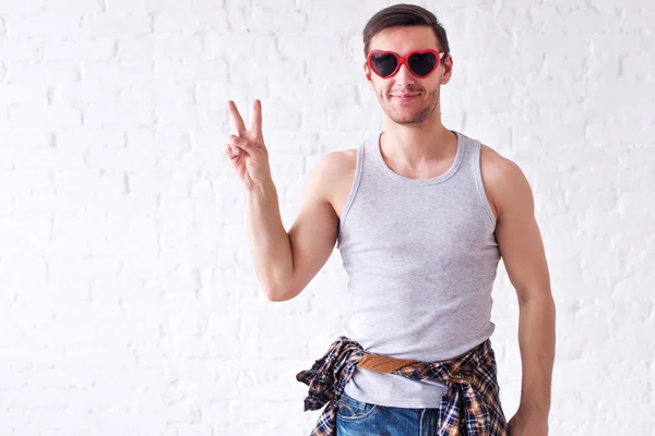 Porträt eines lustig lächelnden Hipster-Mannes mit roter herzförmiger Brille, der in die Kamera blickt und das Friedenszeichen oder die Siegesgeste zeigt — Stockfoto