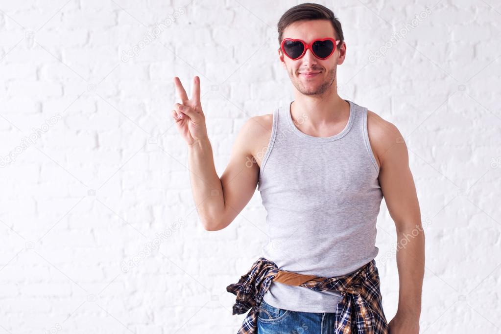 Portrait of funny smiling hipster man in red heart shaped glasses looking at camera showing peace v-sign or victory gesture casual summer urban style
