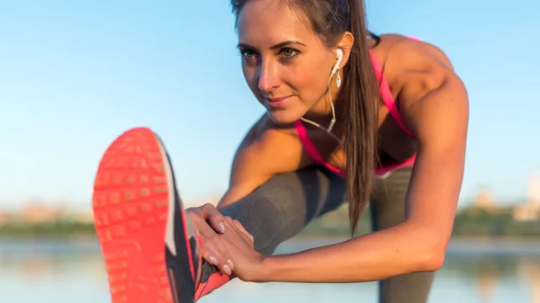 Modelo de fitness atleta menina aquecer esticando seus isquiotibiais, perna e costas. Jovem se exercitando com fones de ouvido ouvindo música ao ar livre na praia ou campo de esportes à noite de verão — Fotografia de Stock