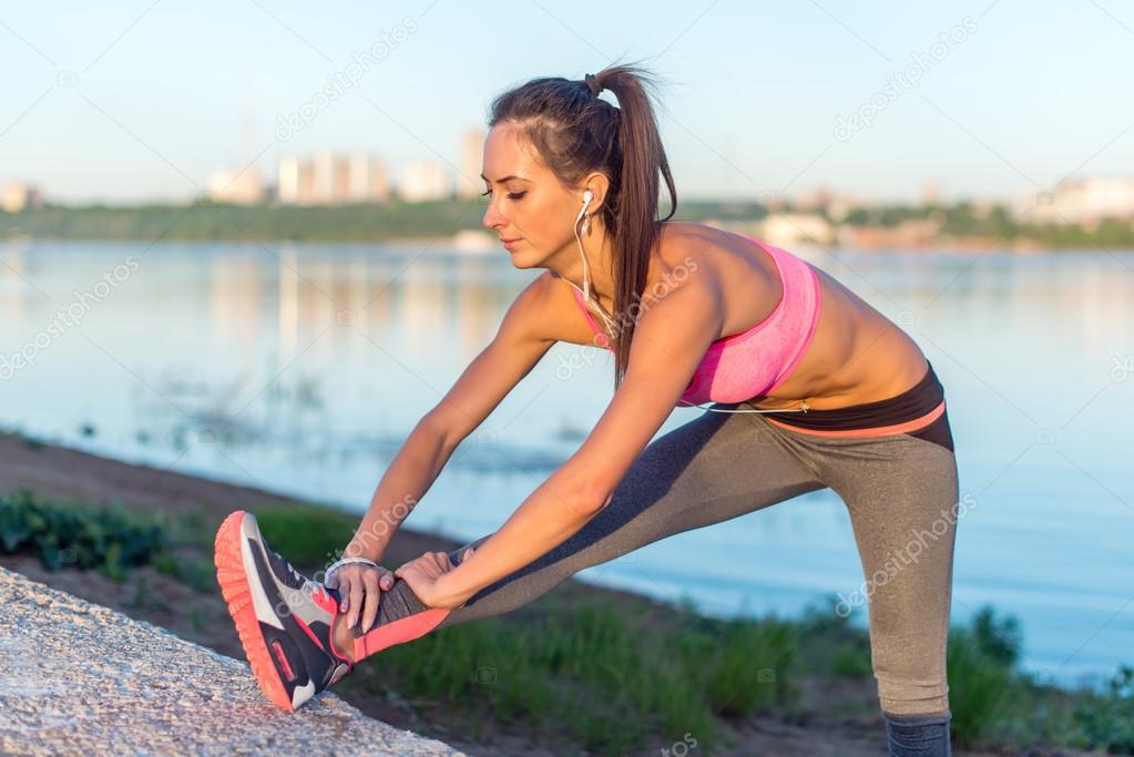 Fitness model athlete girl warm up stretching her hamstrings, leg and back. Young woman exercising with headphones listening music outdoors on beach or sports ground at evening summer