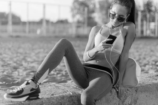 Beautiful fitness athlete woman wearing sunglasses resting listening music after work out exercising on summer evening in beach at sunset with smartphone outdoor portrait. — Stock Photo, Image