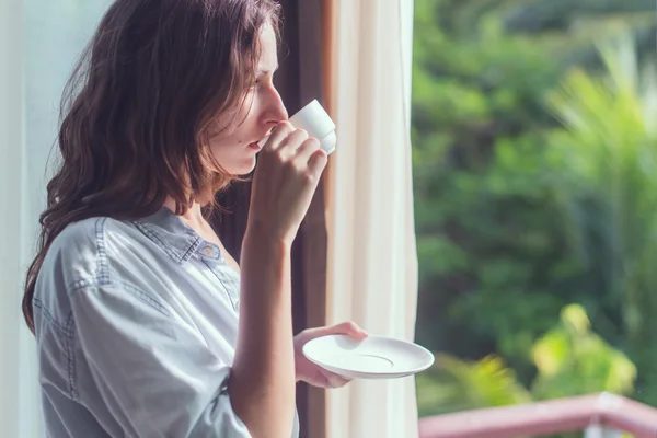 Mulher a beber chá ou café e a olhar pela janela. Jovem senhora reunião nascer do sol . — Fotografia de Stock