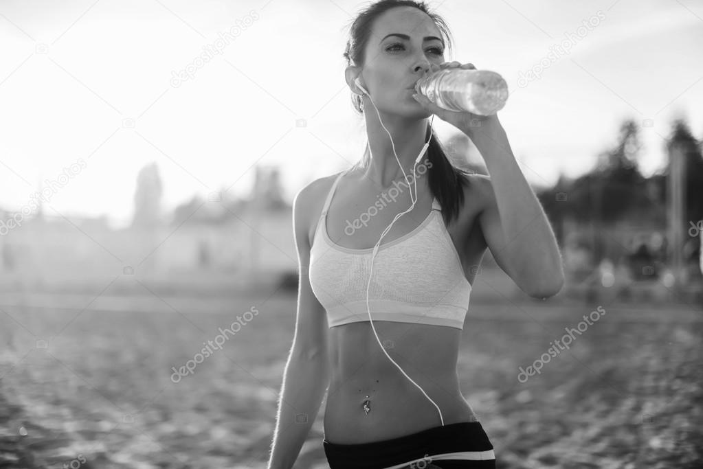 Beautiful fitness athlete woman resting drinking water after work out exercising on beach summer evening in sunny sunshine outdoor portrait