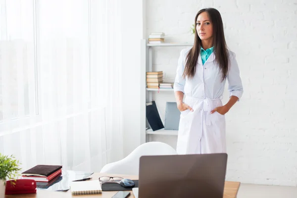 Portret van jonge vrouw arts met een witte vacht staande in medische kantoor kijken naar de camera. — Stockfoto