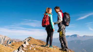 Bir çift, açık bir günde, nefes kesici manzaralarla çevriliyken, bir dağın zirvesinde birbirlerine gülümserler..