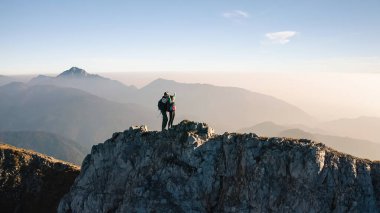 İki yürüyüşçü, gün batımında açık mavi bir gökyüzünün altındaki engin dağ sıralarına hayran kalarak kayalık bir zirvenin tepesinde duruyor..